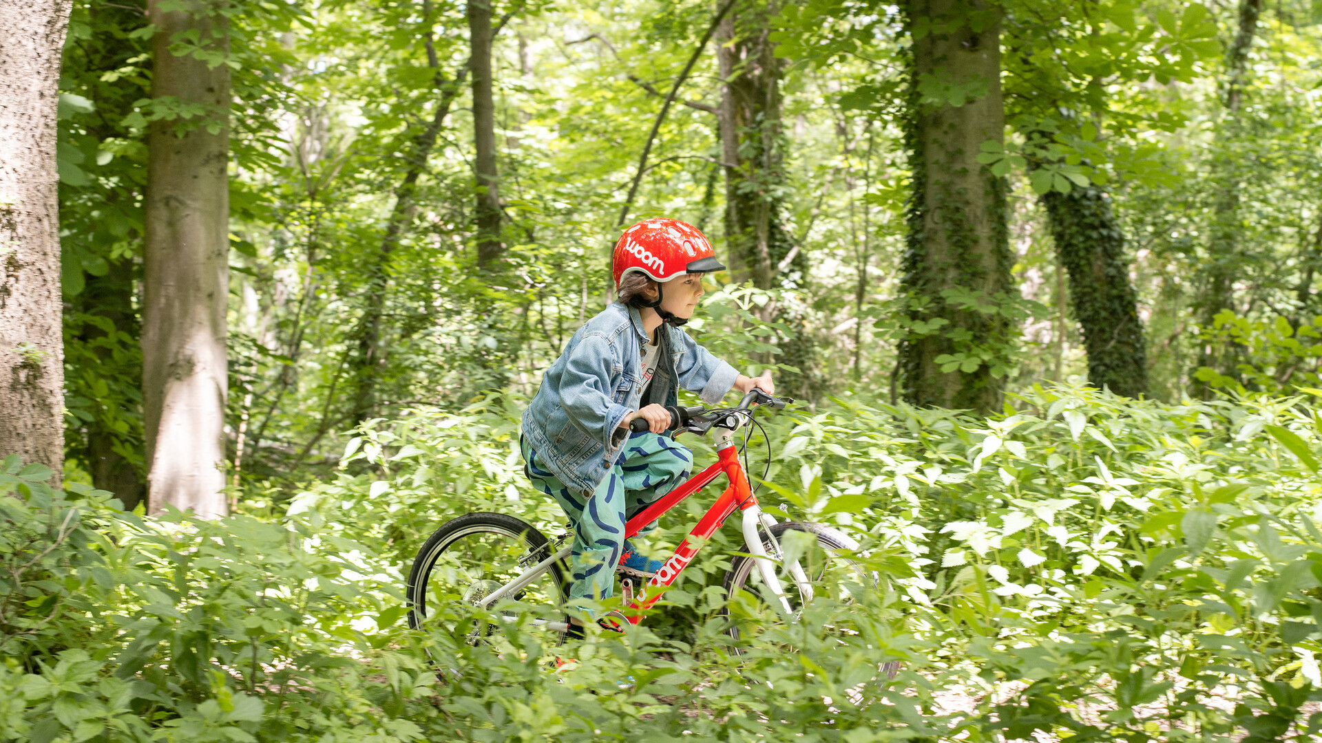 Kind mit rotem Fahrradhelm fährt auf einem roten woom bike durch den Wald.