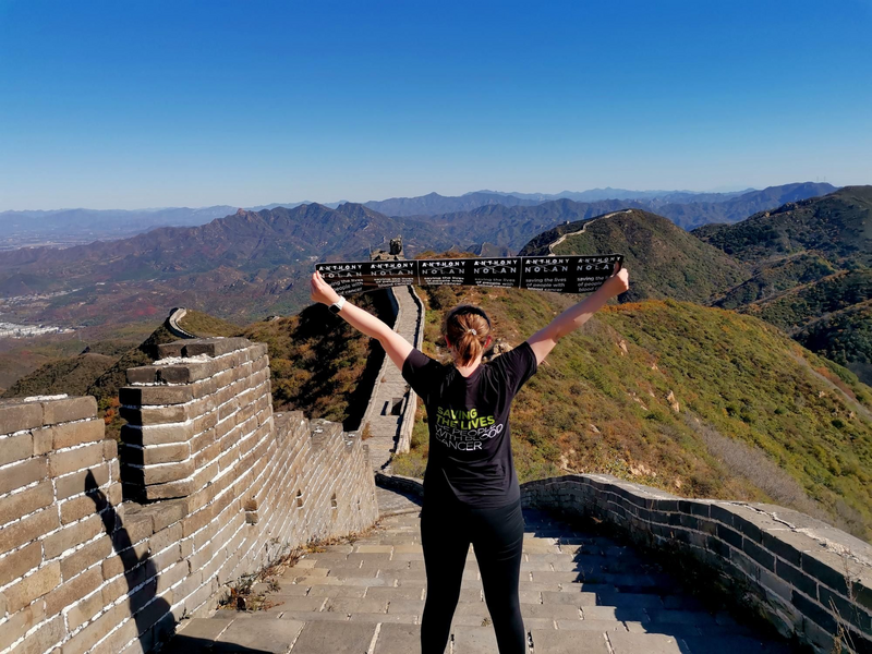 Photo of a woman with an Anthony Nolan tshirt on and banner, standing on the Great Wall of China