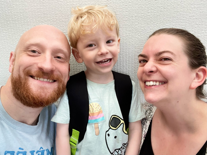 a smiling 3 year old blonde boy stands in the middle of his parents, mum on the right, dad on the left