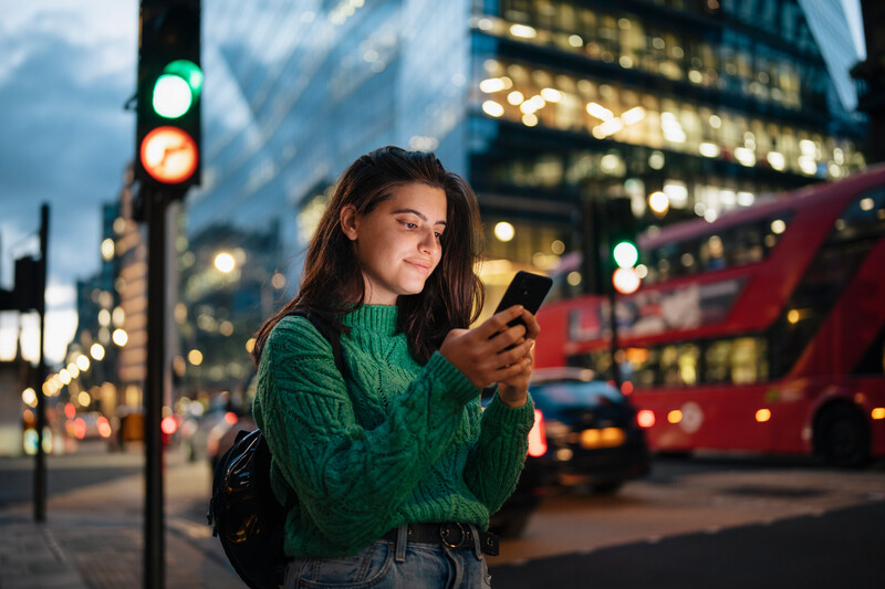 Smiling young woman using phone in the city streets at night.