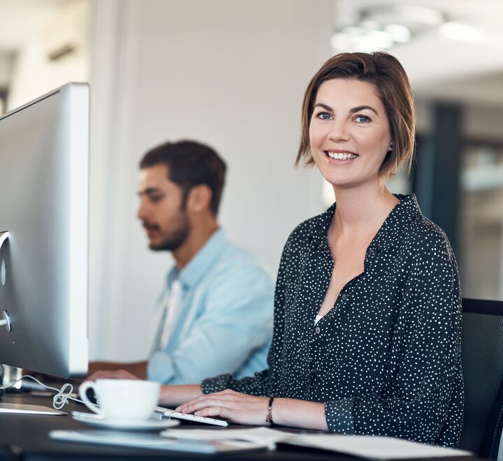 Bilde av en kvinne og en mann som jobber foran hver sin PC. Damen smiler til kamera.