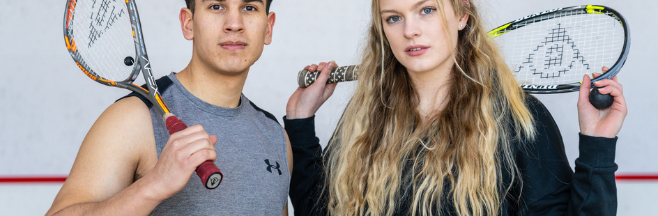 Een jonge vrouw en een jonge man kijken recht in de camera en houden een squashracket vast.