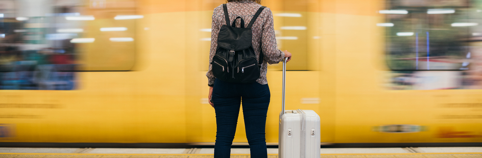 Vrouw met rolkoffer wacht  op perron op trein terwijl er een trein voorbijraast