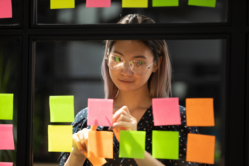 Smiling millennial Asian woman worker busy thinking writing ideas on colorful sticky notes on glass office wall, ethnic female employee brainstorm manage strategy plan project on post it stickers