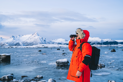 Male traveler with backpack standing against frozen sea with snowy mountains and looking away through binoculars exploring nature in Norway
