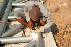 A welder is sitting on a steel roof frame and is welding steel pipes and wearing masks and leather gloves.
