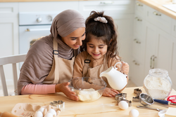 Young Muslim Mom Baking Together With Her Little Daughter In Kitchen. Cute Little Arab Girl Helping Mom To Prepare Dough, Adding Ingredients To Bowl, Islamic Family Having Fun While Cooking At Home