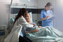 African doctor specialist using stethoscope listening senior man heart, laying in bed breathing with help from oxygen tube, and medical nurse is taking notes on clipboard.