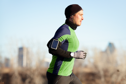 Running man jogging in autumn listening to music on smart phone. Runner training in warm outfit on cold day. Fit male fitness athlete model training outdoor in fall. Full body length of jogger.