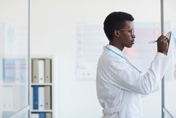 Side view portrait of young African-American doctor writing on clipboard while standing by glass wall in med clinic, copy space