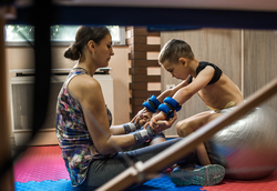 Physical therapist assisting little boy make exercises on gym ball during rehabilitation in children hospital.