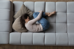 Frustrated depressed millennial girl lying in embryo pose on couch, sleeping, drowsing, feeling stress, apathy, suffering from abdominal pain, bellyache. Depression concept. Top view
