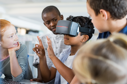 Mixed race child with vr virtual reality goggles in classroom. Multiethnic pupil having fun with virtual reality headset at elementary school. Happy boy gesturing while using VR headset in classroom.