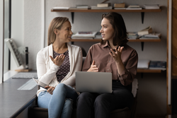 Two motivated business women chatting, discussing online project ideas or working issues using computer software applications, sitting in modern office room, professional cooperation concept.