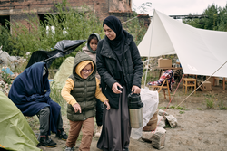 Muslim mother in hijab carrying thermos flask along refugee camp while her daughter running around her playfully